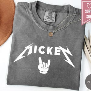 Pu&ograve; includere: Una maglietta grigia con la scritta "MICKEY" in bianco, con un carattere stilizzato, e un gesto della mano bianco. La maglietta ha uno scollo rotondo e maniche corte. Una collana d'argento con un ciondolo &egrave; sulla maglietta.