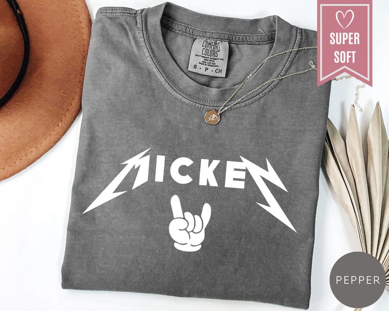 Pu&ograve; includere: Una maglietta grigia con la scritta "MICKEY" in bianco, con un carattere stilizzato, e un gesto della mano bianco. La maglietta ha uno scollo rotondo e maniche corte. Una collana d'argento con un ciondolo &egrave; sulla maglietta.
