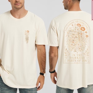 May include: A cream-colored short-sleeve t-shirt featuring a small floral graphic on the front left chest. The back displays a large illustration of a ribcage filled with flowers and the text: FIND SOMEONE WHO GROWS FLOWERS IN THE DARKEST PARTS OF YOU.