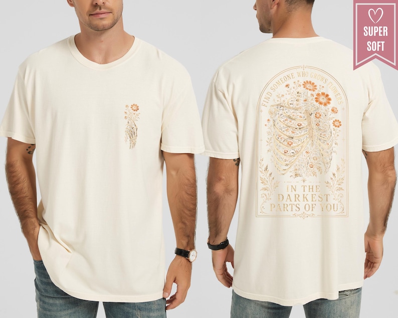 May include: A cream-colored short-sleeve t-shirt featuring a small floral graphic on the front left chest. The back displays a large illustration of a ribcage filled with flowers and the text: FIND SOMEONE WHO GROWS FLOWERS IN THE DARKEST PARTS OF YOU.
