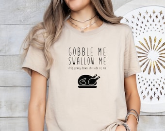 May include: A beige t-shirt with the text "Gobble Me Swallow Me" and "drip gravy down the side of me" in black. A black silhouette of a roasted turkey is below the text.