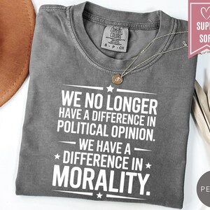May include: A gray t-shirt with the text "WE NO LONGER HAVE A DIFFERENCE IN POLITICAL OPINION. WE HAVE A DIFFERENCE IN MORALITY." A silver necklace with a pendant is on the shirt. The shirt is next to a brown hat.
