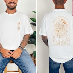 May include: A white t-shirt featuring a small floral graphic on the front left chest. The back displays a large arched design of a ribcage intertwined with flowers and the text: FIND SOMEONE WHO GROWS FLOWERS IN THE DARKEST PARTS OF YOU.