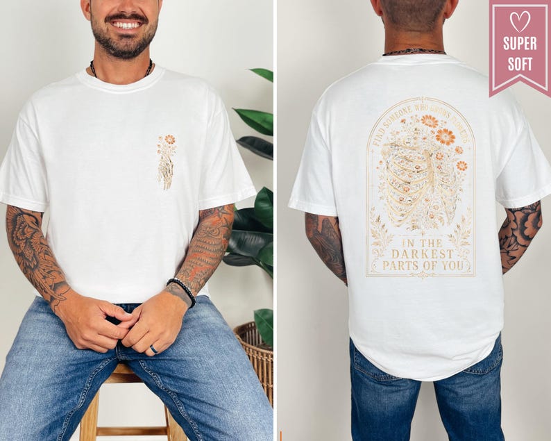 May include: A white t-shirt featuring a small floral graphic on the front left chest. The back displays a large arched design of a ribcage intertwined with flowers and the text: FIND SOMEONE WHO GROWS FLOWERS IN THE DARKEST PARTS OF YOU.