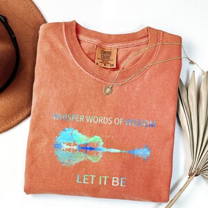 Puede incluir: Una camiseta Comfort Colors de color coral con las palabras "Whisper Words of Wisdom" y "Let It Be" impresas en letras iridiscentes. Un gr&aacute;fico de guitarra aparece debajo del texto. Un collar de oro y un sombrero marr&oacute;n tambi&eacute;n est&aacute;n en la imagen.