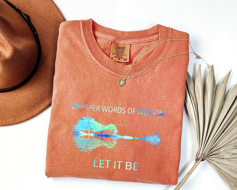 Puede incluir: Una camiseta Comfort Colors de color coral con las palabras "Whisper Words of Wisdom" y "Let It Be" impresas en letras iridiscentes. Un gr&aacute;fico de guitarra aparece debajo del texto. Un collar de oro y un sombrero marr&oacute;n tambi&eacute;n est&aacute;n en la imagen.