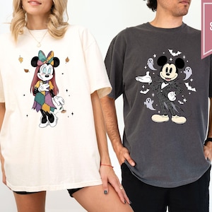 Puede incluir: Camisetas color marfil y pimienta con personajes de Disney. La camiseta marfil muestra a Minnie Mouse con un vestido de retazos, mientras que la camiseta pimienta muestra a Mickey Mouse con un atuendo inspirado en Jack Skellington, rodeado de fantasmas y murci&eacute;lagos.