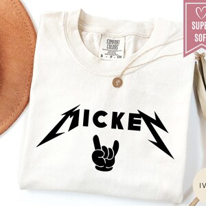 Pu&ograve; includere: T-shirt color avorio con la scritta "MICKEY" in un carattere nero stilizzato e una grafica di gesto della mano nera. La maglietta ha uno scollo tondo e una piccola etichetta in alto a sinistra. &Egrave; visibile una collana d'oro.