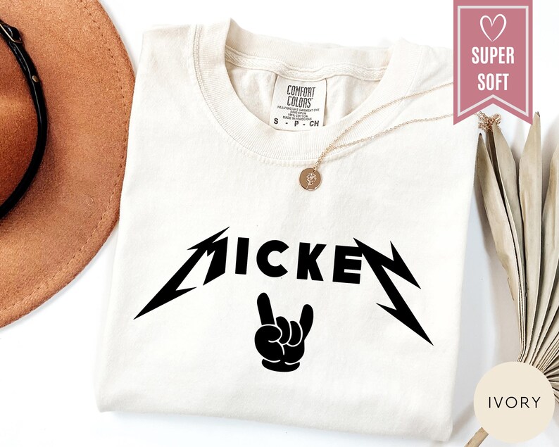 Pu&ograve; includere: T-shirt color avorio con la scritta "MICKEY" in un carattere nero stilizzato e una grafica di gesto della mano nera. La maglietta ha uno scollo tondo e una piccola etichetta in alto a sinistra. &Egrave; visibile una collana d'oro.