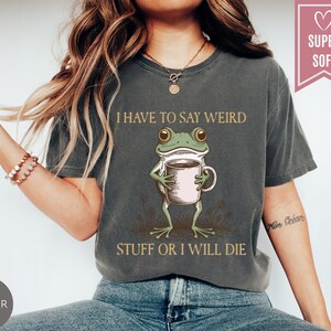 May include: Dark gray t-shirt with a graphic of a frog holding a coffee cup. The text reads "I HAVE TO SAY WEIRD STUFF OR I WILL DIE." A pink heart-shaped tag says "SUPER SOFT."