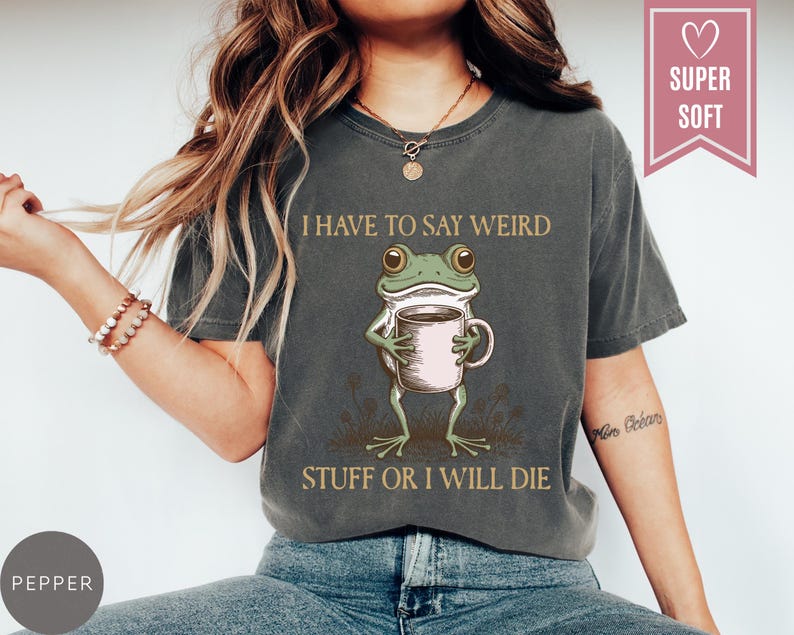May include: Dark gray t-shirt with a graphic of a frog holding a coffee cup. The text reads "I HAVE TO SAY WEIRD STUFF OR I WILL DIE." A pink heart-shaped tag says "SUPER SOFT."