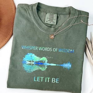 Puede incluir: Camiseta verde oliva con las palabras "Whisper Words of Wisdom" y "Let It Be" impresas en azul hologr&aacute;fico. La camiseta presenta un gr&aacute;fico de guitarra con un dise&ntilde;o de reflejo de &aacute;rbol y agua. Tambi&eacute;n se ven un collar dorado y un sombrero marr&oacute;n.
