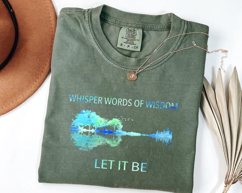 Puede incluir: Camiseta verde oliva con las palabras "Whisper Words of Wisdom" y "Let It Be" impresas en azul hologr&aacute;fico. La camiseta presenta un gr&aacute;fico de guitarra con un dise&ntilde;o de reflejo de &aacute;rbol y agua. Tambi&eacute;n se ven un collar dorado y un sombrero marr&oacute;n.