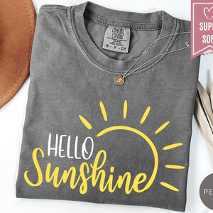 Puede incluir: Una camiseta gris con las palabras "Hello Sunshine" en blanco y amarillo, con un gr&aacute;fico de sol amarillo. La camiseta tiene cuello redondo y mangas cortas. Un collar plateado con un colgante est&aacute; sobre la camiseta.
