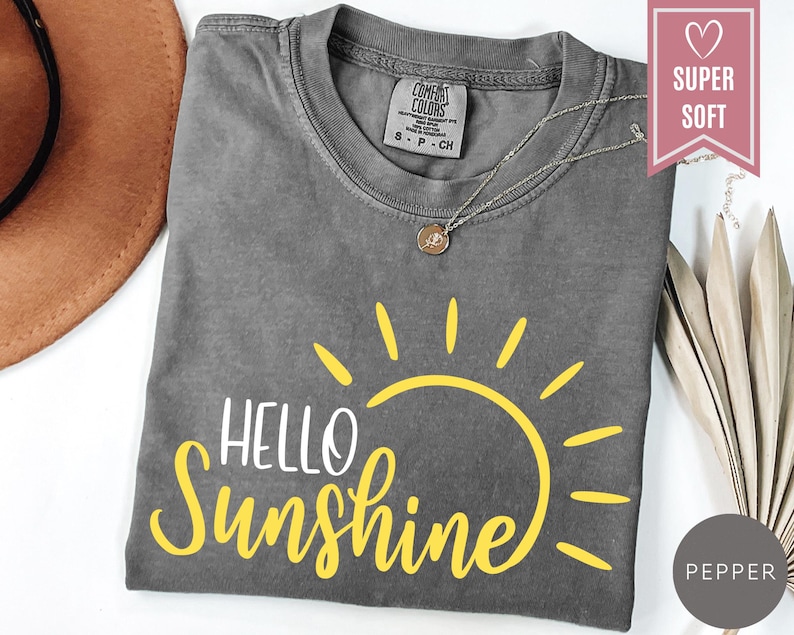 Puede incluir: Una camiseta gris con las palabras "Hello Sunshine" en blanco y amarillo, con un gr&aacute;fico de sol amarillo. La camiseta tiene cuello redondo y mangas cortas. Un collar plateado con un colgante est&aacute; sobre la camiseta.