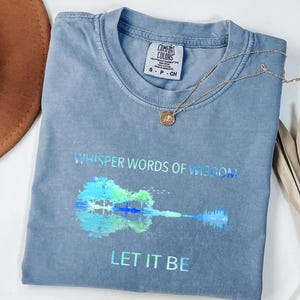 Puede incluir: Una camiseta azul Comfort Colors con las palabras "Whisper Words of Wisdom" y "Let It Be" impresas. Un gr&aacute;fico de guitarra colorida est&aacute; debajo del texto. Un collar dorado con un colgante descansa sobre la camisa.