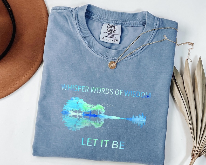 Puede incluir: Una camiseta azul Comfort Colors con las palabras "Whisper Words of Wisdom" y "Let It Be" impresas. Un gr&aacute;fico de guitarra colorida est&aacute; debajo del texto. Un collar dorado con un colgante descansa sobre la camisa.