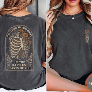May include: A charcoal gray t-shirt featuring a graphic on the back of a human ribcage intertwined with orange flowers and the text "FIND SOMEONE WHO GROWS FLOWERS IN THE DARKEST PARTS OF YOU." The front features a small graphic of a skeletal hand.
