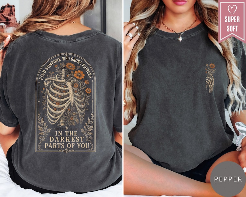 May include: A charcoal gray t-shirt featuring a graphic on the back of a human ribcage intertwined with orange flowers and the text "FIND SOMEONE WHO GROWS FLOWERS IN THE DARKEST PARTS OF YOU." The front features a small graphic of a skeletal hand.