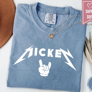 Pu&ograve; includere: Una maglietta blu jeans con la scritta "MICKEY" in un carattere bianco stilizzato, con un gesto della mano rock and roll sotto. Una collana d'oro con un ciondolo &egrave; drappeggiata sulla maglietta.