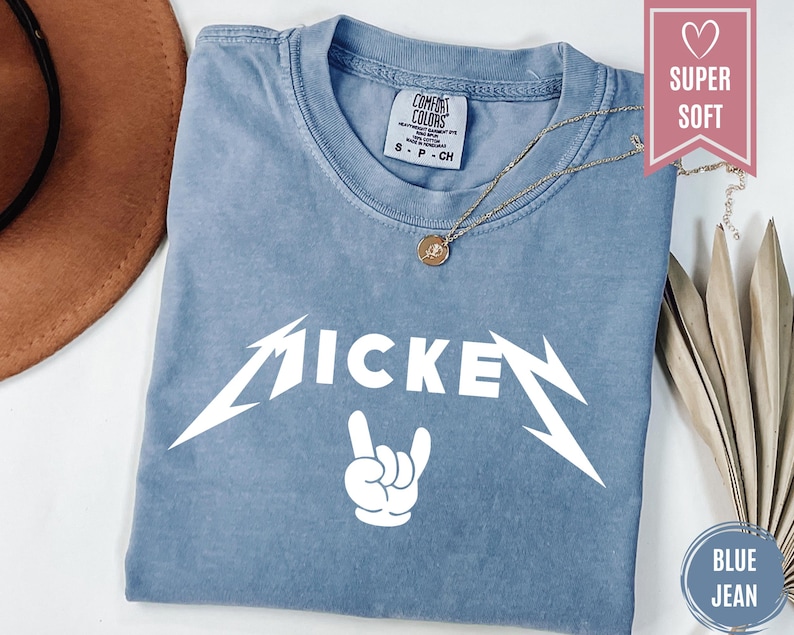 Pu&ograve; includere: Una maglietta blu jeans con la scritta "MICKEY" in un carattere bianco stilizzato, con un gesto della mano rock and roll sotto. Una collana d'oro con un ciondolo &egrave; drappeggiata sulla maglietta.