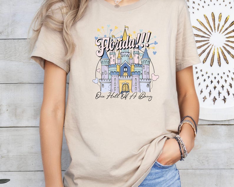 May include: A beige t-shirt with a colorful illustration of a castle and the text "Florida!!! One Hell of a  Dream".