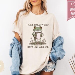 May include: Ivory-colored t-shirt featuring a whimsical frog holding a coffee cup. The frog is illustrated with green and brown details, standing amidst small plants. The text "I HAVE TO SAY WEIRD STUFF OR I WILL DIE" is printed above the frog. A pink banner reads "SUPER SOFT".