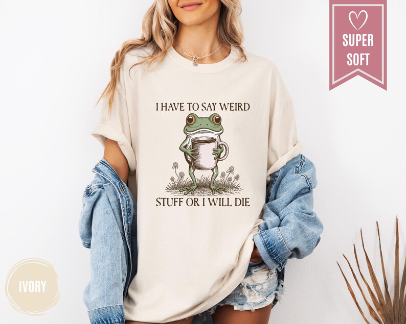 May include: Ivory-colored t-shirt featuring a whimsical frog holding a coffee cup. The frog is illustrated with green and brown details, standing amidst small plants. The text "I HAVE TO SAY WEIRD STUFF OR I WILL DIE" is printed above the frog. A pink banner reads "SUPER SOFT".