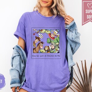 Op de afbeelding: Violet T-shirt met een afbeelding van Toy Story-personages, waaronder Woody, Buzz Lightyear en de aliens, in een zwart kader. De tekst "YOU'VE GOT A FRIEND IN ME" staat eronder. Het shirt heeft een "SUPER SOFT" label.