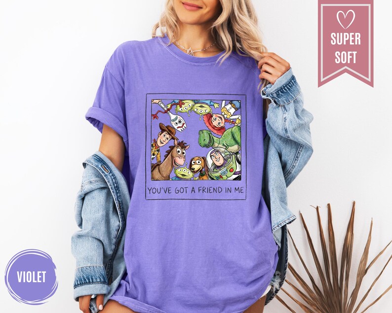 Op de afbeelding: Violet T-shirt met een afbeelding van Toy Story-personages, waaronder Woody, Buzz Lightyear en de aliens, in een zwart kader. De tekst "YOU'VE GOT A FRIEND IN ME" staat eronder. Het shirt heeft een "SUPER SOFT" label.