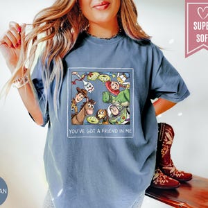 Op de afbeelding: Blauwe jeanskleurige t-shirt met een kleurrijke afbeelding van personages uit een populaire animatiefilm. De afbeelding is omlijst in een witte rechthoek en bevat de tekst "YOU'VE GOT A FRIEND IN ME". Een roze hartvormig label leest "SUPER SOFT".
