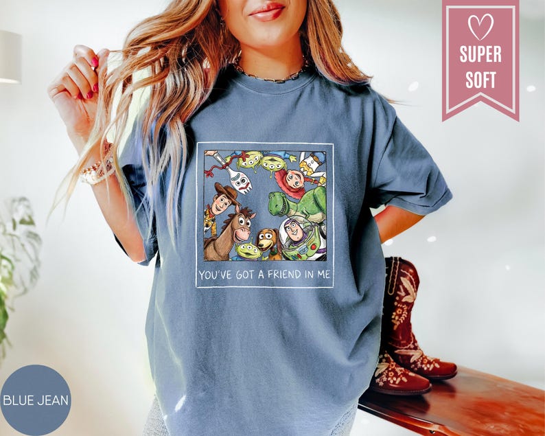 Op de afbeelding: Blauwe jeanskleurige t-shirt met een kleurrijke afbeelding van personages uit een populaire animatiefilm. De afbeelding is omlijst in een witte rechthoek en bevat de tekst "YOU'VE GOT A FRIEND IN ME". Een roze hartvormig label leest "SUPER SOFT".