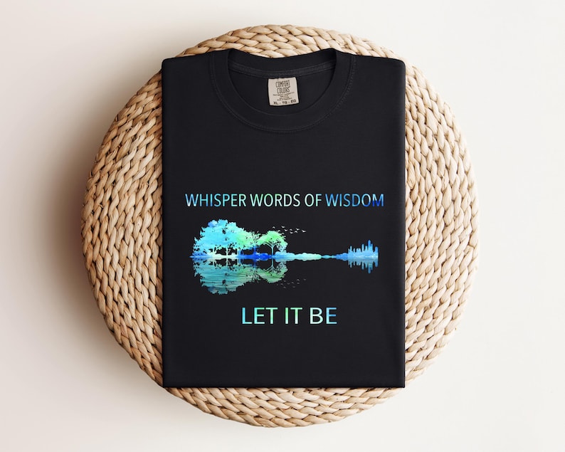 Puede incluir: Camiseta negra con el texto "WHISPER WORDS OF WISDOM" sobre un dise&ntilde;o en forma de guitarra con un &aacute;rbol y un reflejo de agua, y las palabras "LET IT BE" debajo. El dise&ntilde;o utiliza tonos azules y verdes.