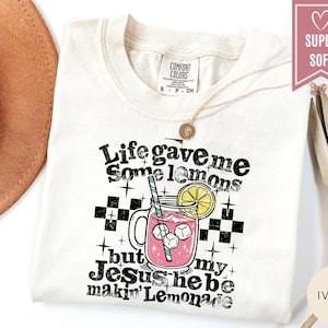 May include: Ivory-colored t-shirt with the text "Life gave me some lemons but Jesus he be makin' Lemonade" in a distressed font. The graphic includes a pink lemonade drink with ice cubes and a lemon slice. A gold necklace is visible.