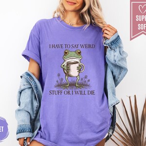 May include: Violet-colored t-shirt with the text "I HAVE TO SAY WEIRD STUFF OR I WILL DIE." The graphic features a cartoon frog holding a mug. The shirt is labeled "SUPER SOFT."