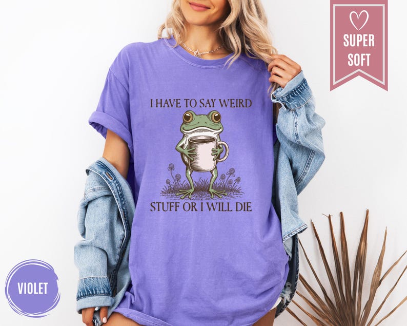 May include: Violet-colored t-shirt with the text "I HAVE TO SAY WEIRD STUFF OR I WILL DIE." The graphic features a cartoon frog holding a mug. The shirt is labeled "SUPER SOFT."