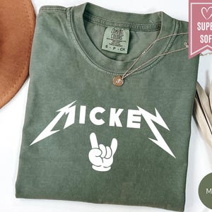 Pu&ograve; includere: Una maglietta verde muschio con la scritta "MICKEY" in bianco, con un carattere stilizzato, e un gesto della mano bianco. La maglietta ha un girocollo e maniche corte. Una collana d'oro con un ciondolo &egrave; sulla maglietta.