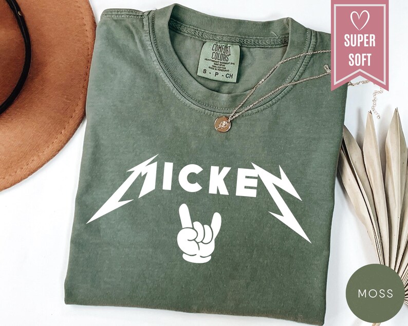 Pu&ograve; includere: Una maglietta verde muschio con la scritta "MICKEY" in bianco, con un carattere stilizzato, e un gesto della mano bianco. La maglietta ha un girocollo e maniche corte. Una collana d'oro con un ciondolo &egrave; sulla maglietta.