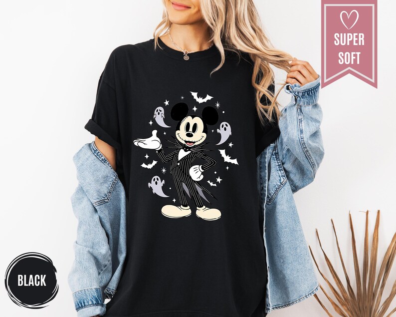 Puede incluir: Camiseta negra con un dise&ntilde;o de Halloween de Mickey Mouse como Jack Skellington, rodeado de murci&eacute;lagos y fantasmas. La camiseta tiene el texto "SUPER SOFT" en una pancarta rosa y "BLACK" en un c&iacute;rculo negro.