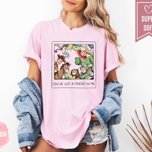 Op de afbeelding: Lichtroze T-shirt met een ingelijste afbeelding van Toy Story-personages, waaronder Woody en Buzz Lightyear. De tekst "YOU'VE GOT A FRIEND IN ME" staat eronder. Het T-shirt heeft opgerolde mouwen. Het woord "SUPER SOFT" staat in een roze banner.