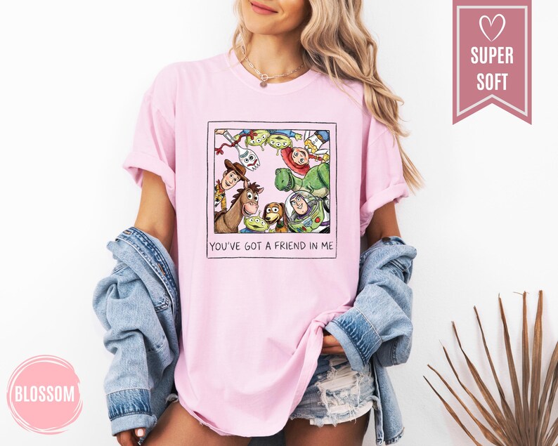 Op de afbeelding: Lichtroze T-shirt met een ingelijste afbeelding van Toy Story-personages, waaronder Woody en Buzz Lightyear. De tekst "YOU'VE GOT A FRIEND IN ME" staat eronder. Het T-shirt heeft opgerolde mouwen. Het woord "SUPER SOFT" staat in een roze banner.