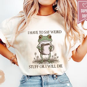 May include: Ivory-colored t-shirt featuring a whimsical illustration of a green frog holding a coffee mug. The text above the frog reads "I HAVE TO SAY WEIRD" and below it, "STUFF OR I WILL DIE". The shirt has a "SUPER SOFT" label.