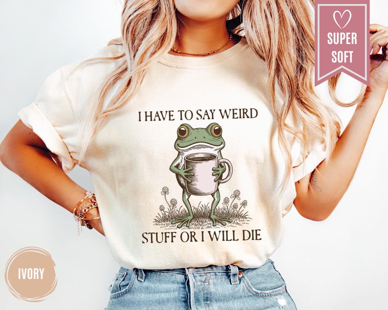 May include: Ivory-colored t-shirt featuring a whimsical illustration of a green frog holding a coffee mug. The text above the frog reads "I HAVE TO SAY WEIRD" and below it, "STUFF OR I WILL DIE". The shirt has a "SUPER SOFT" label.