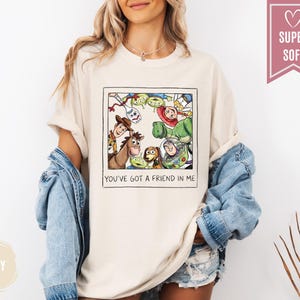 Op de afbeelding: Ivoor kleurig T-shirt met een ingelijste afbeelding van Toy Story-personages, waaronder Woody, Buzz Lightyear en de Little Green Men. De tekst "YOU'VE GOT A FRIEND IN ME" staat eronder. Het shirt heeft korte mouwen.