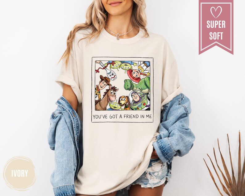 Op de afbeelding: Ivoor kleurig T-shirt met een ingelijste afbeelding van Toy Story-personages, waaronder Woody, Buzz Lightyear en de Little Green Men. De tekst "YOU'VE GOT A FRIEND IN ME" staat eronder. Het shirt heeft korte mouwen.
