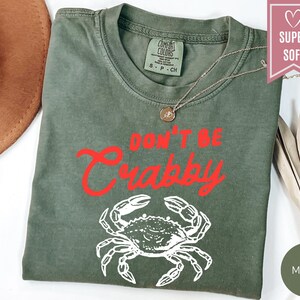 May include: Moss green t-shirt with the phrase "DON'T BE CRABBY" in red script and a white crab graphic. The shirt has a crew neck and short sleeves. A gold necklace is draped on the shirt.