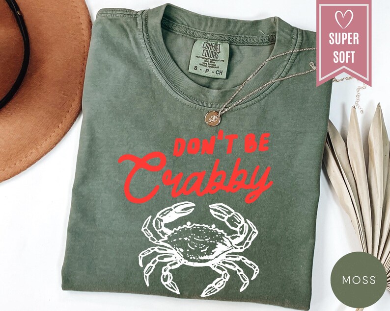 May include: Moss green t-shirt with the phrase "DON'T BE CRABBY" in red script and a white crab graphic. The shirt has a crew neck and short sleeves. A gold necklace is draped on the shirt.