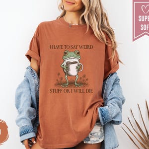 May include: A rust-colored t-shirt with the text "I HAVE TO SAY WEIRD STUFF OR I WILL DIE" and a cartoon frog holding a coffee cup. The shirt is a relaxed fit and has rolled-up sleeves.