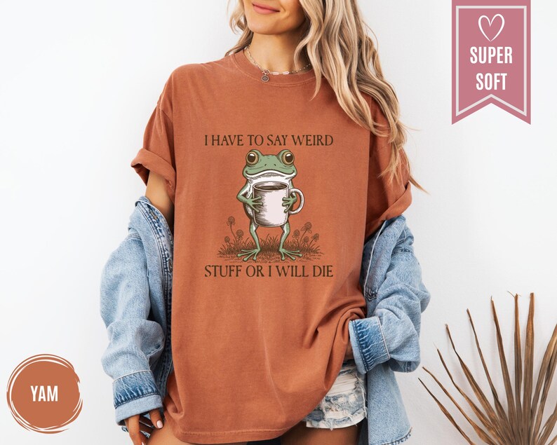 May include: A rust-colored t-shirt with the text "I HAVE TO SAY WEIRD STUFF OR I WILL DIE" and a cartoon frog holding a coffee cup. The shirt is a relaxed fit and has rolled-up sleeves.