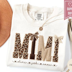 May include: Ivory-colored t-shirt with the word "MIMI" in a leopard print design. The letters are accented with brown and beige details and bows. Below the word are the names "olivia", "josh", and "emma". A gold necklace is visible.
